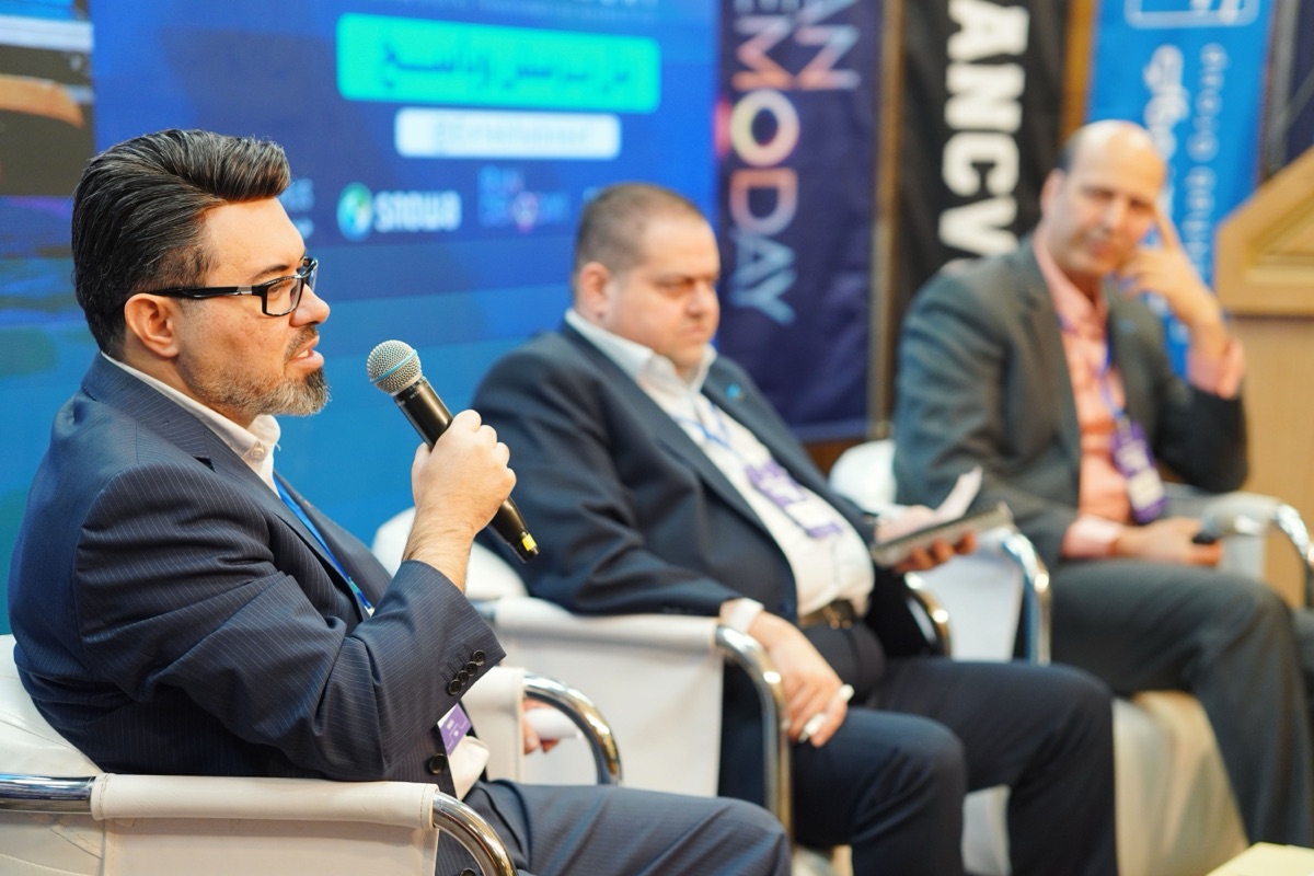 A panel of speakers seated on stage at a business forum, one addressing the audience through a microphone