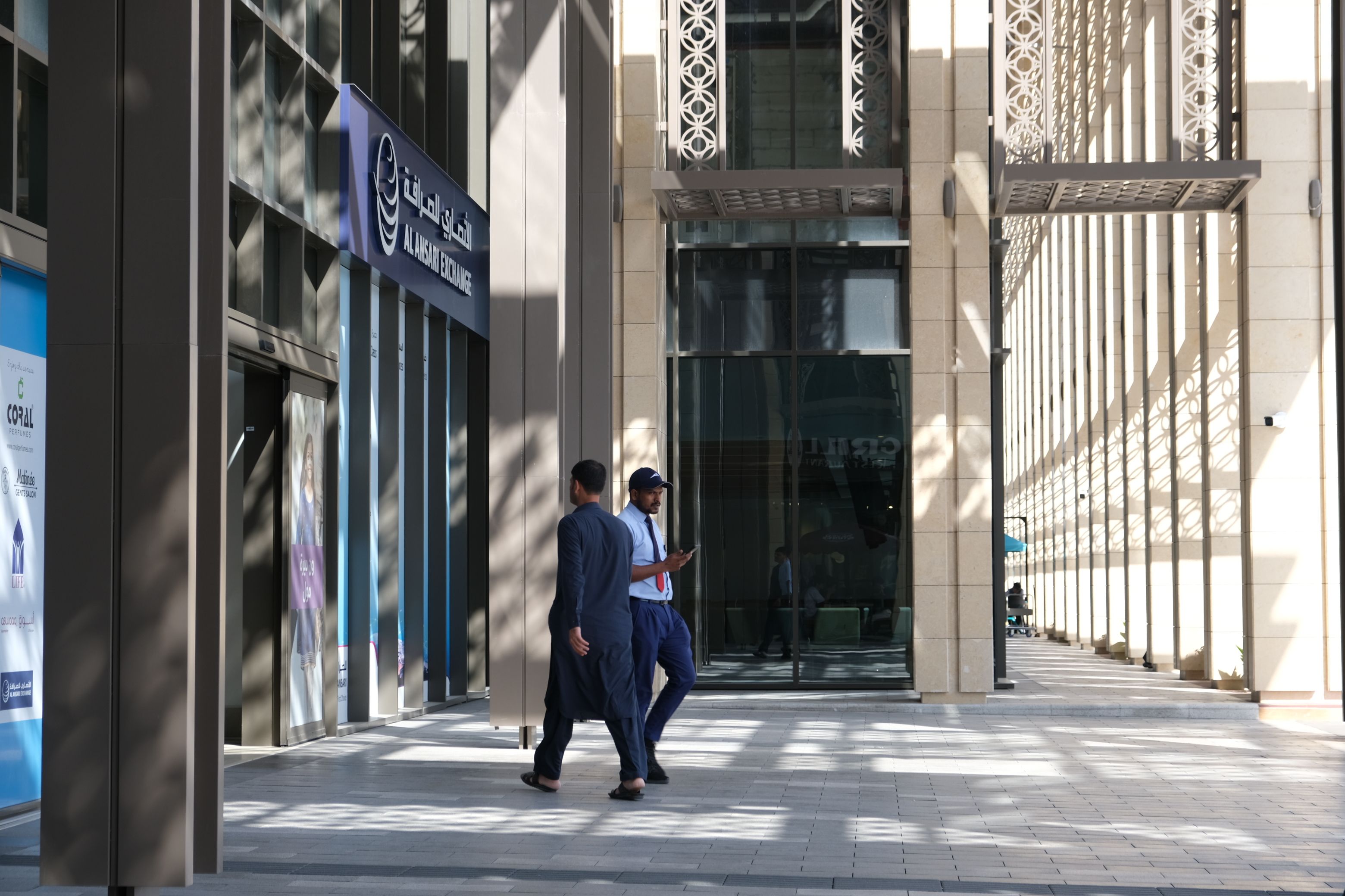 Pedestrians walking past an Al Ansari Exchange branch in a sunlit ground floor retail colonnade of a modern UAE office tower